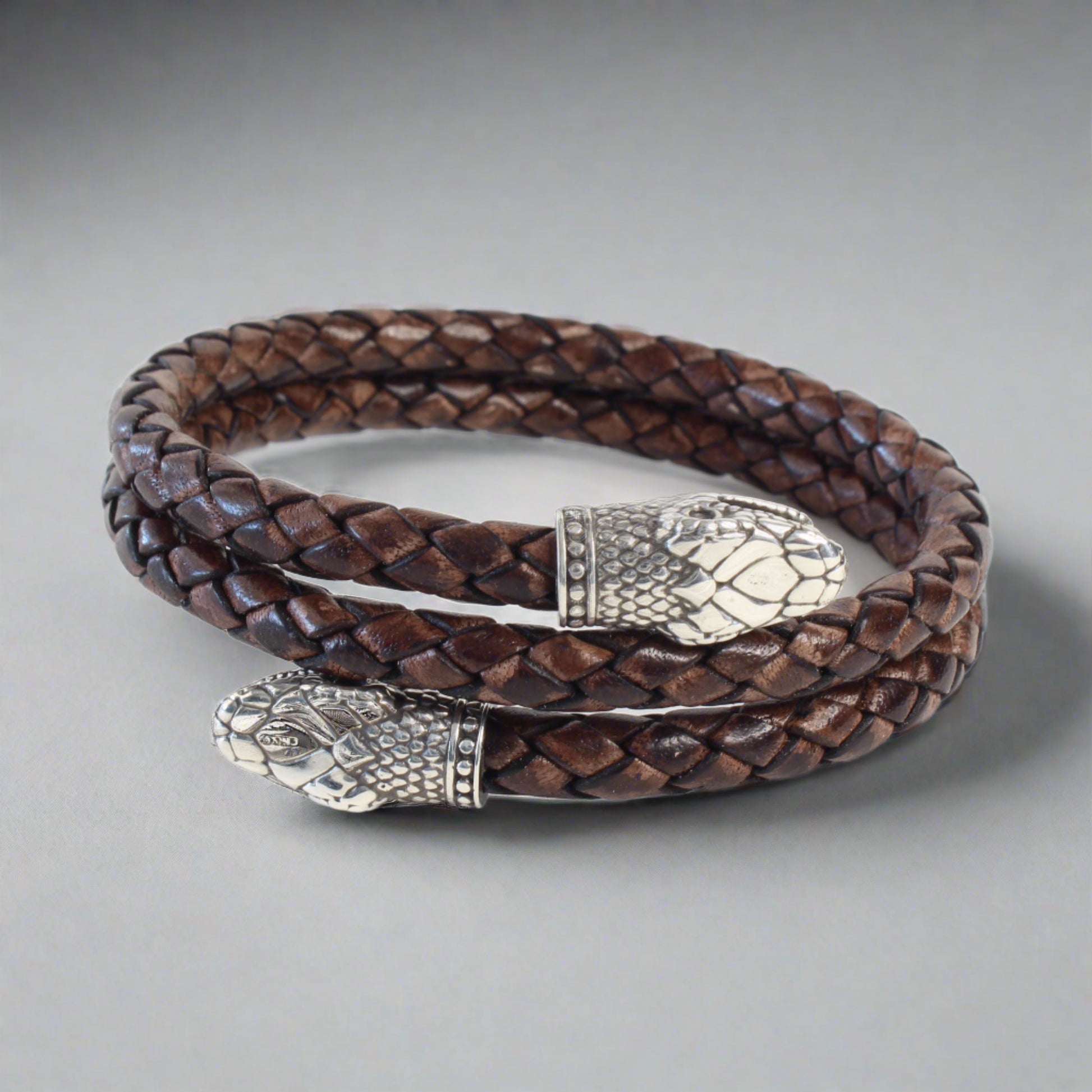 Handcrafted braided leather bracelet in dark brown, coiled in a circle with two silver snake head end pieces featuring detailed scales and facial features. The design blends rugged texture with intricate metalwork, highlighting artisanal craftsmanship.