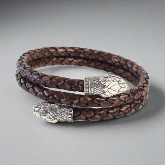 Handcrafted braided leather bracelet in dark brown, coiled in a circle with two silver snake head end pieces featuring detailed scales and facial features. The design blends rugged texture with intricate metalwork, highlighting artisanal craftsmanship.