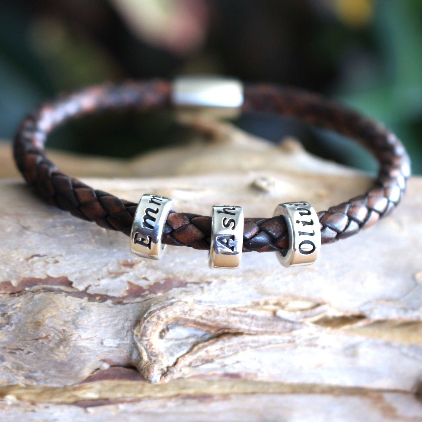 Braided antique brown leather bracelet with three sterling silver engraved name beads reading Emma, Asher, and Olivia, finished with a secure metallic clasp.