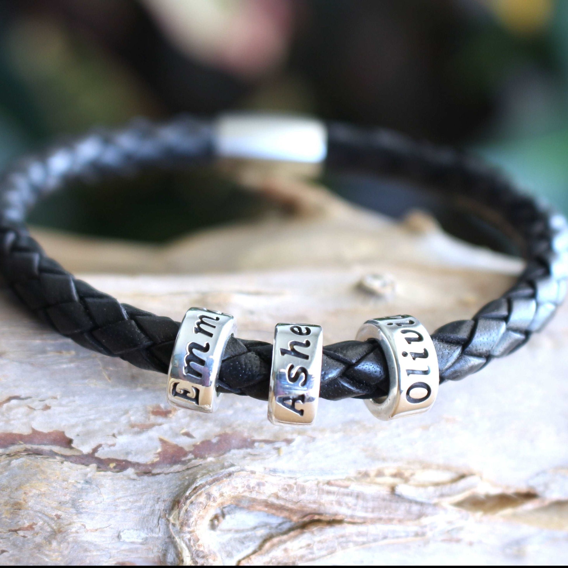 Braided black leather bracelet with three sterling silver engraved name beads reading Emma, Asher, and Olivia, finished with a secure metallic clasp.