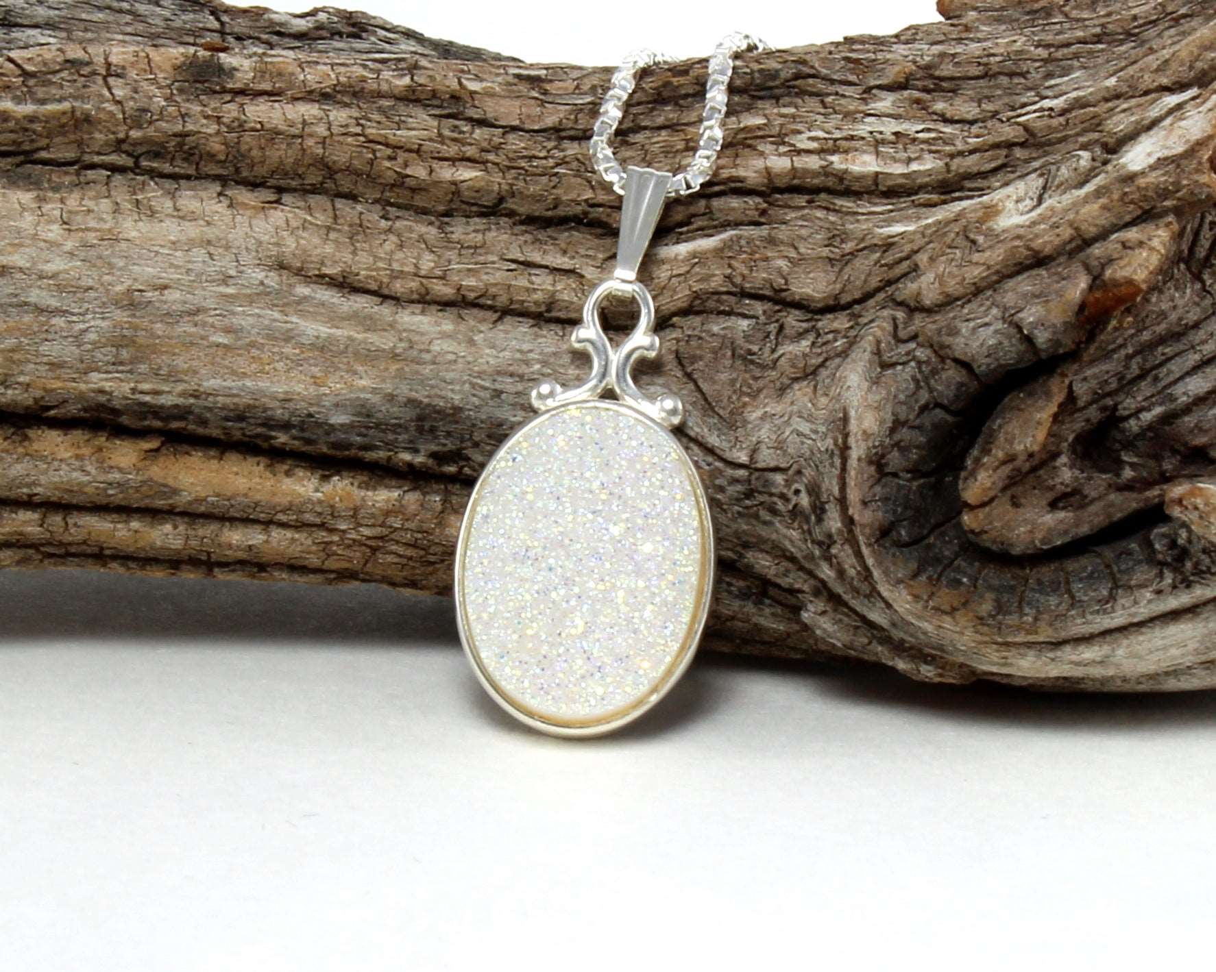 Oval silver druzy pendant necklace with glittery white stone on a textured silver chain, displayed against rustic wood background.
