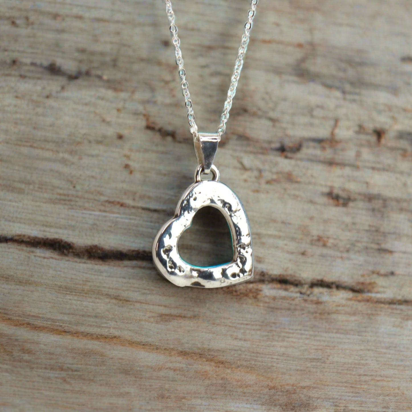 Silver heart-shaped pendant with a hollow center and textured edges, displayed on a rustic wooden surface with a delicate silver chain.