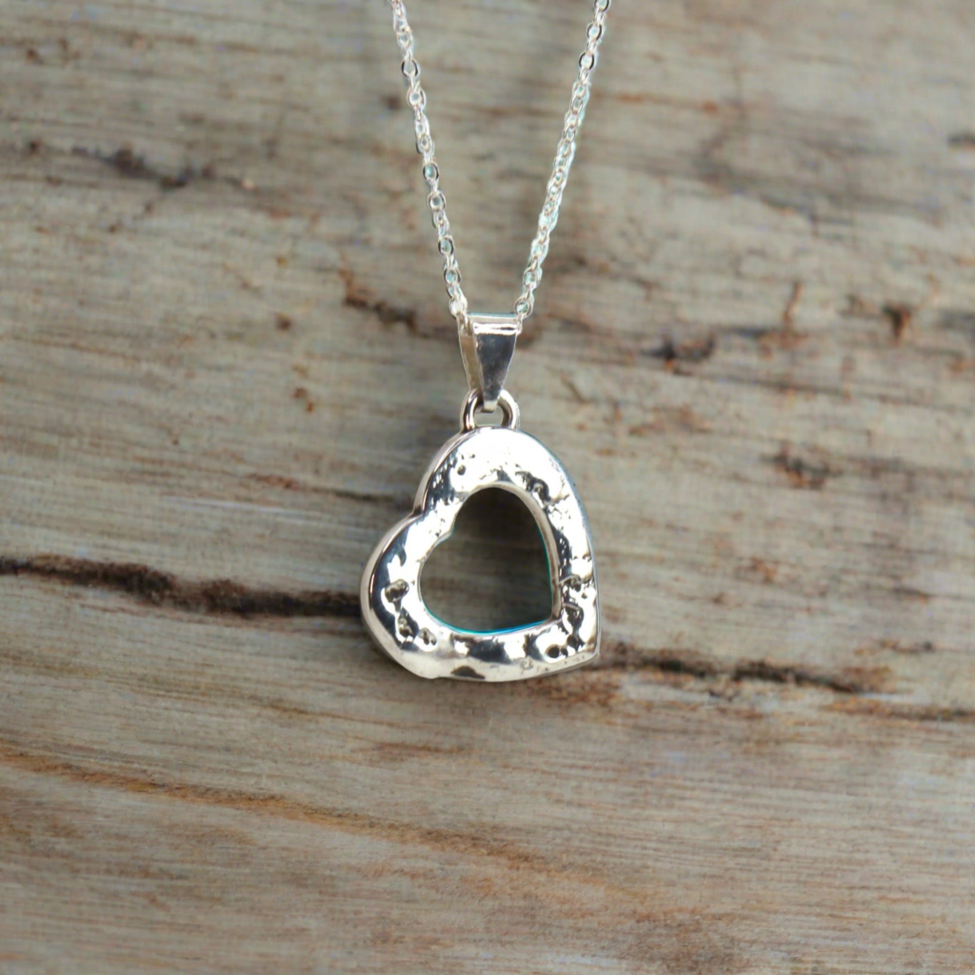 Silver heart-shaped pendant with a hollow center and textured edges, displayed on a rustic wooden surface with a delicate silver chain.