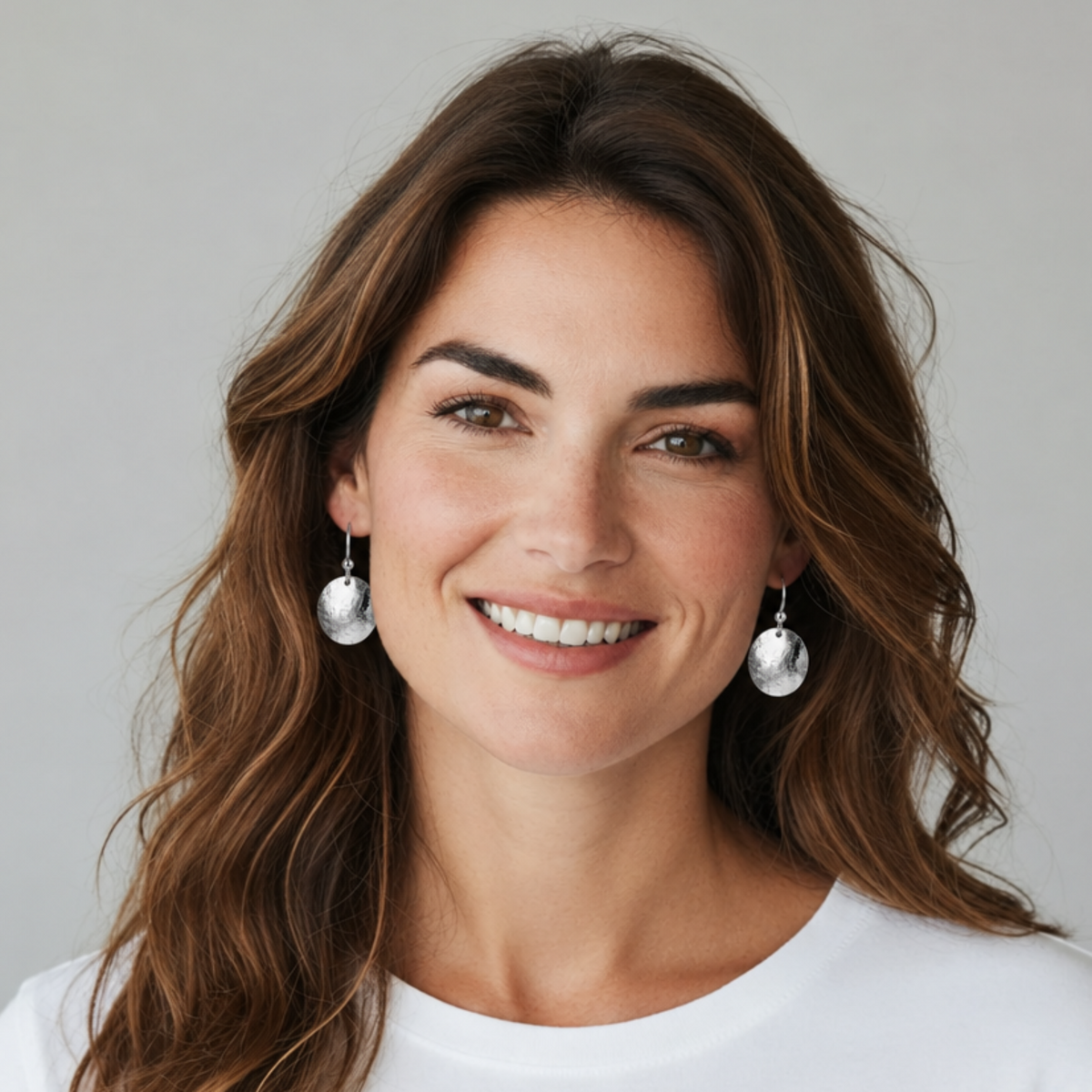 A smiling woman with long wavy brown hair wearing silver dangle earrings with white circular drops, photographed against a light gray background.