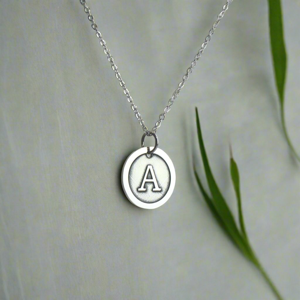 Sterling silver necklace with a circular pendant featuring a raised letter "A" in the center, displayed on soft fabric with green leaves for a natural accent.